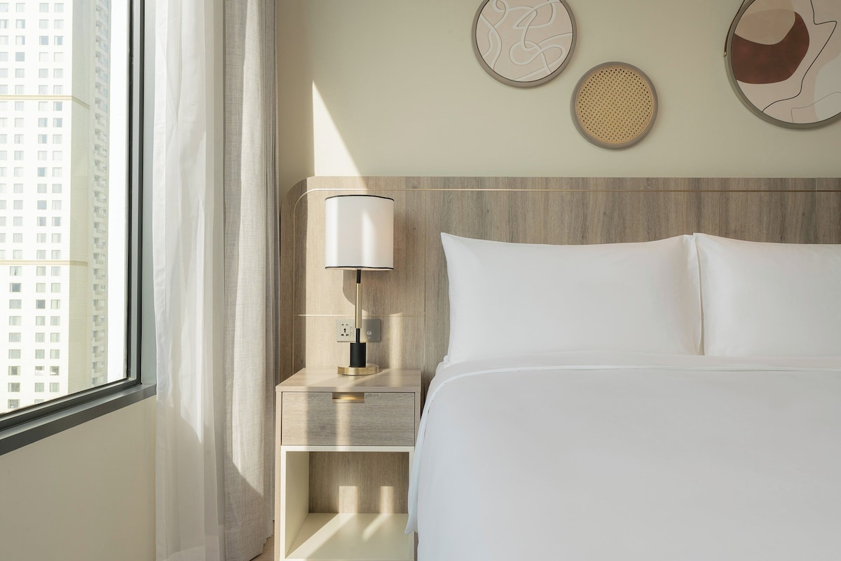 Une chambre d’hôtel avec un lit, une table de chevet avec une lampe et une fenêtre donnant sur des immeubles.