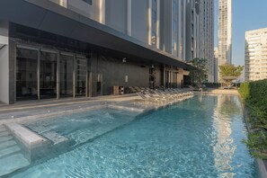 Una piscina con sillones de descanso, un edificio moderno con amplios ventanales y un cielo despejado.