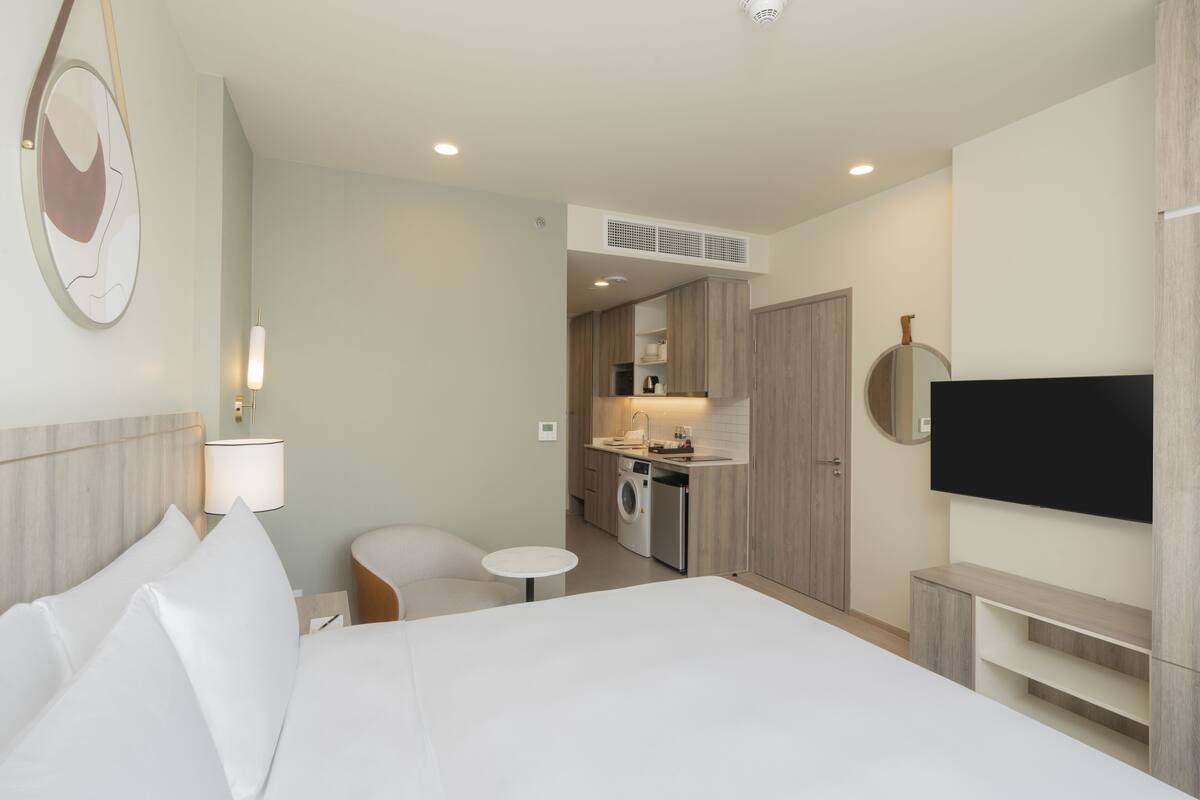Une chambre d’hôtel moderne avec un grand lit, une petite table ronde, un canapé, un téléviseur fixé au mur et, au fond, une kitchenette.