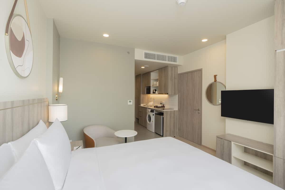 Una habitación de hotel moderna con una cama grande, una pequeña mesa redonda, un sofá, un televisor montado en la pared y una kitchenette al fondo.