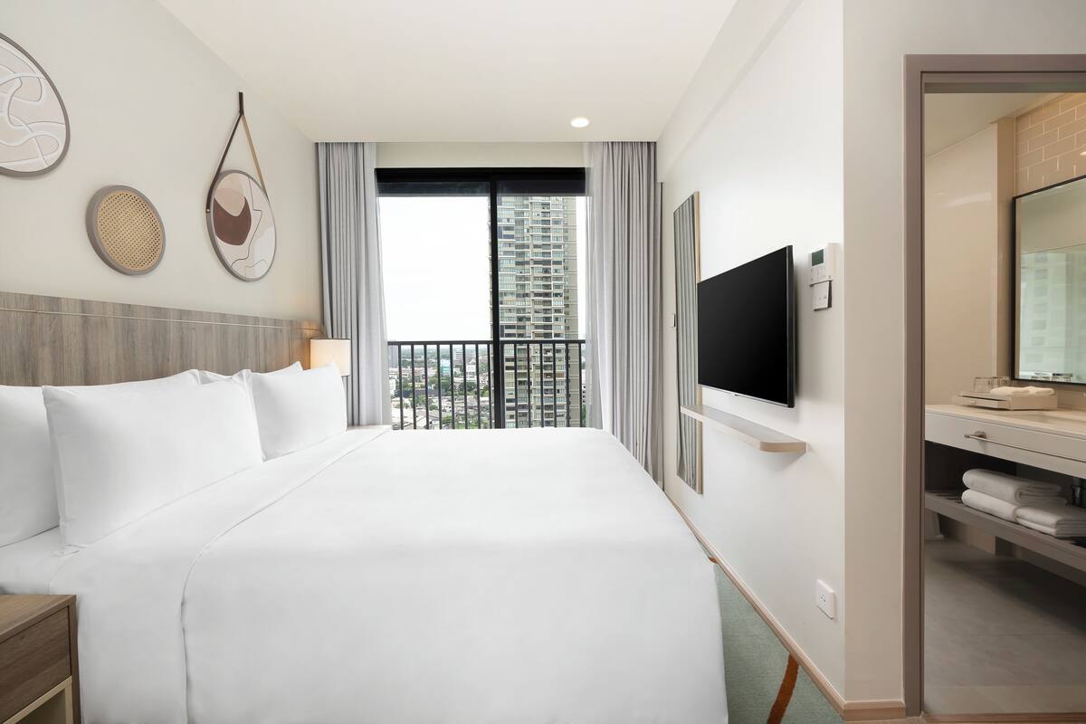 Une chambre d’hôtel avec un grand lit, un téléviseur fixé au mur, un balcon avec vue sur la ville et une salle de bain équipée d’un miroir et d’un porte-serviettes.
