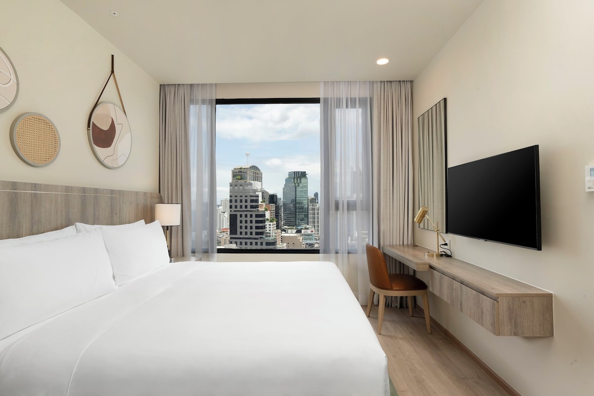 Une chambre d’hôtel avec un grand lit, un bureau avec une chaise, un téléviseur fixé au mur et une vue sur le paysage urbain à travers de fines rideaux.