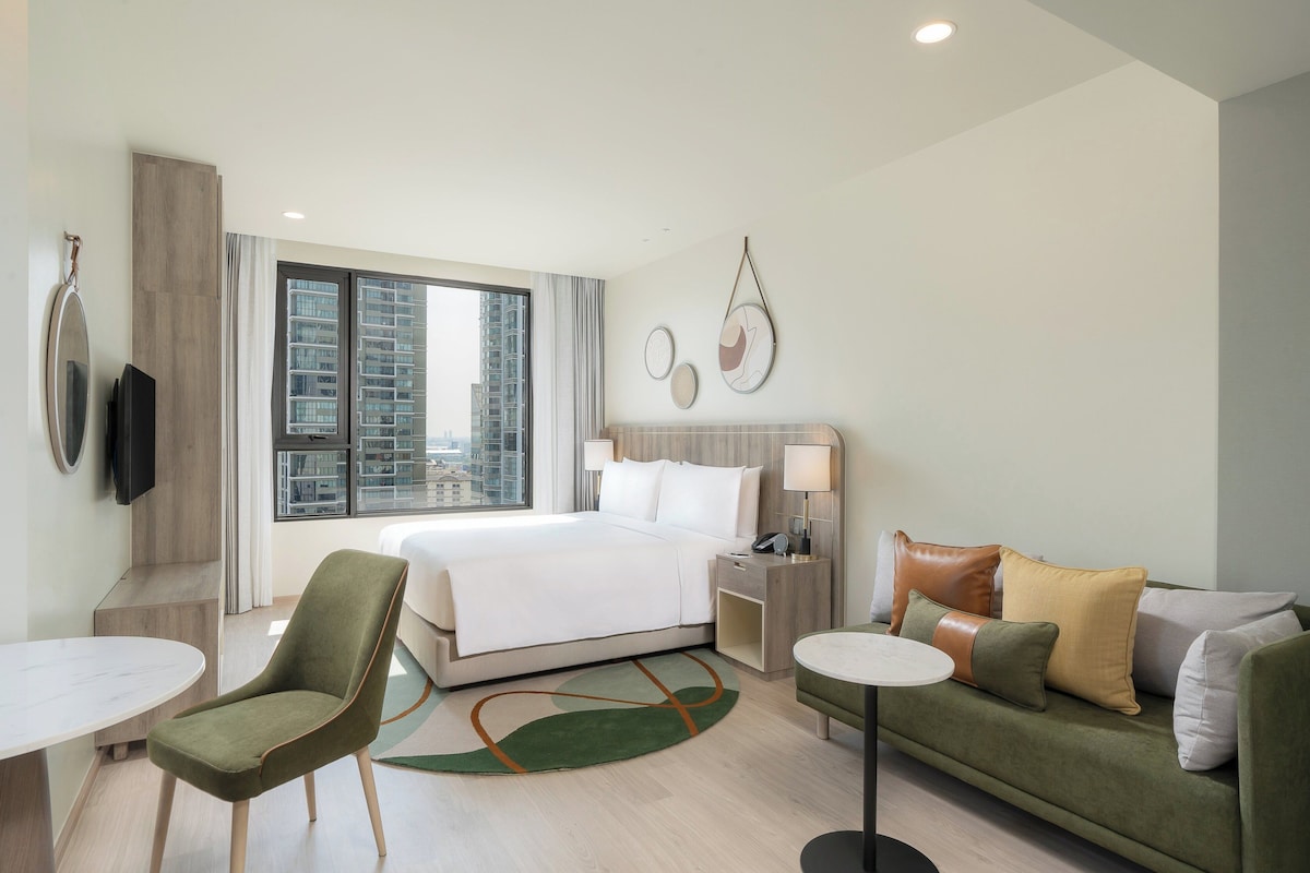 Une chambre d’hôtel moderne avec un grand lit, un fauteuil vert, un canapé, une table ronde et un téléviseur.