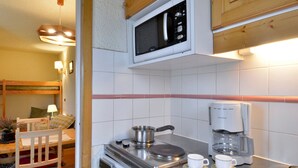 A compact kitchen with a microwave, coffee maker, and a small stove. There is a wooden dining table with chairs and a bunk bed in the background. - Cozy Studio 4 pers · Ski-in · Pets Allowed (Belle Plagne)