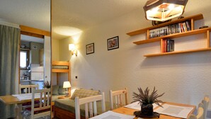 A cozy room with a wooden dining table, two chairs, a bench, a small kitchen area, and a bookshelf with books and decorative items. - Cozy Studio 4 pers · Ski-in · Pets Allowed (Belle Plagne)