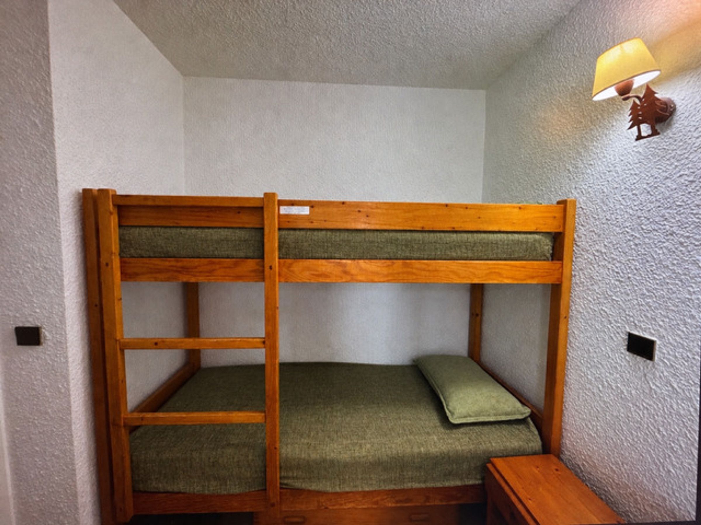 A bunk bed with a wooden frame and green bedding, a small wooden nightstand, and a wall-mounted lamp.