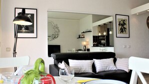 A modern living room with a black sofa, white cushions, a dining table with plates and glasses, and a kitchen area in the background. - Modern Apartment, Sleeps 4, Air-Conditioned with Parking (Villeneuve-Loubet)