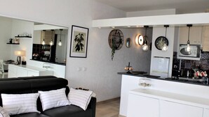A modern living room with a black sofa and a white dining area with a kitchen in the background. - Modern Apartment, Sleeps 4, Air-Conditioned with Parking (Villeneuve-Loubet)