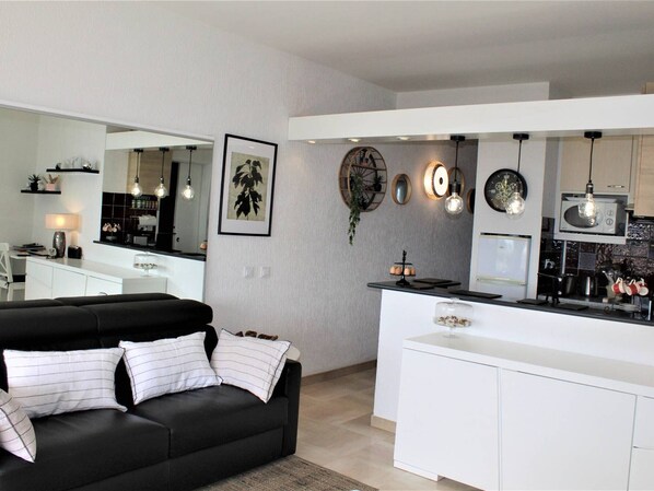 Un salon moderne avec un canapé noir et un coin repas blanc, la cuisine étant visible à l’arrière-plan.
