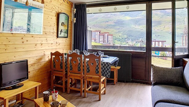 Ein gemütliches Zimmer mit Holzwänden, einem Esstisch mit Stühlen, einem Fernseher und einem großen Fenster mit Blick auf eine gebirgige Landschaft.