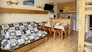 Um quarto aconchegante com sofá, uma mesa de jantar posta para dois, área de cozinha com armários de madeira e uma televisão fixada na parede.