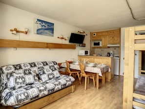 A cozy room with a sofa, a dining table set for two, a kitchen area with wooden cabinets, and a television mounted on the wall.