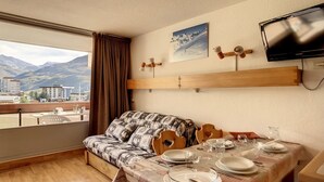 Quarto com cama, mesa de jantar posta com pratos e copos, televisão na parede e janela com vista para montanhas e edifícios.