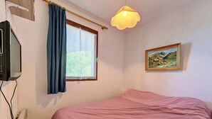 A bedroom with a bed, a television, a window with curtains, a painting, and a hanging light fixture.