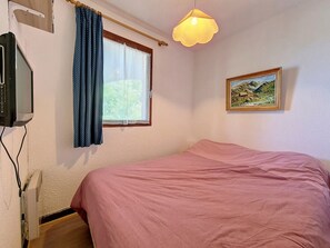 Un dormitorio con cama, televisión, ventana con cortinas, un cuadro y una lámpara colgante.