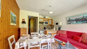 A cozy living room with a red sofa, dining table, and chairs. A kitchen with wooden cabinets and a microwave is visible in the background.