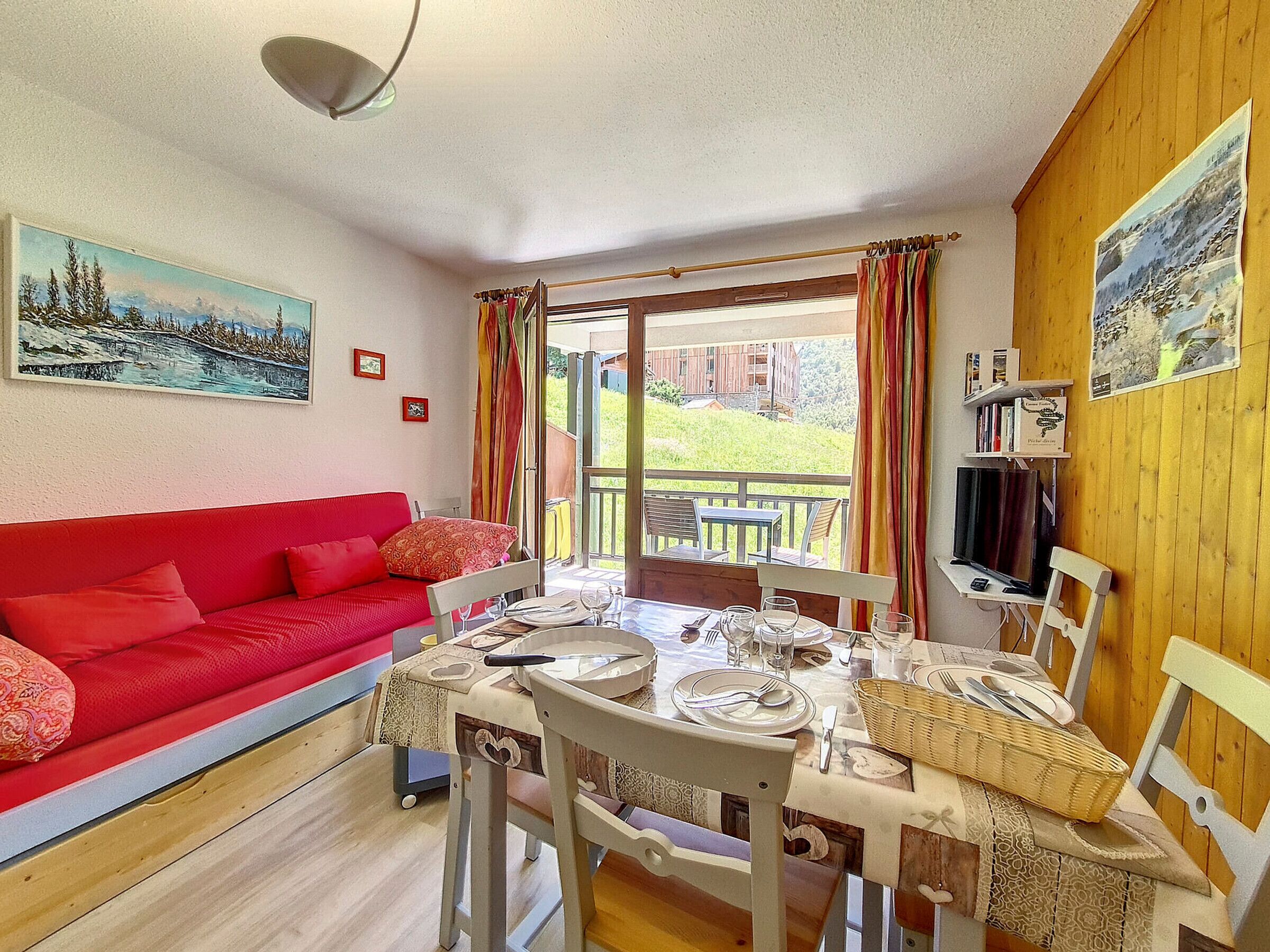 Ein gemütliches Zimmer mit einem roten Sofa, einem Esstisch für vier Personen und Blick auf eine grüne Landschaft durch die Tür.