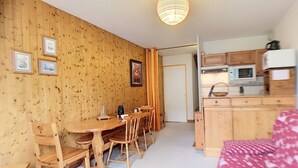 Quarto aconchegante com paredes de madeira, uma mesa de jantar com cadeiras, uma área de cozinha com armários e eletrodomésticos, e uma cama com cobertura estampada.