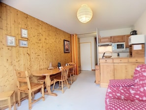A cozy room with wooden walls, a dining table with chairs, a kitchen area with cabinets and appliances, and a bed with a patterned cover.