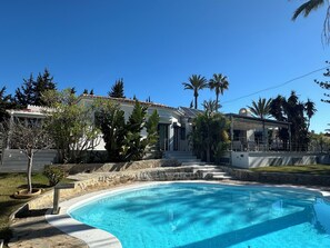 A swimming pool with a stone border, surrounded by a garden with palm trees and a white building with a tiled roof.
