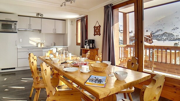 Un comedor con una mesa de madera dispuesta para seis personas, rodeada de sillas de madera. A la izquierda se ve una cocina con armarios blancos y un microondas.