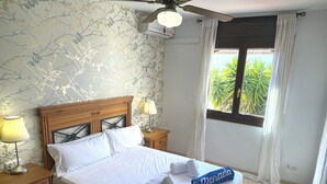 A neatly made bed with white linens and two rolled towels with names on them, a wooden nightstand with a lamp, a ceiling fan, and a window with curtains. - MASIA4 adosado con piscina comunitaria (Miami Playa)