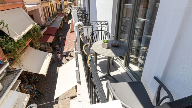 A balcony with a table and chairs, overlooking a street with shops and people. - Metropolitan Pearl Hotel (Chania)