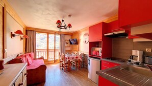 Un cuarto acogedor con piso de madera, una mesa de comedor con sillas, un sofá, una zona de cocina con gabinetes rojos y un televisor montado en la pared.