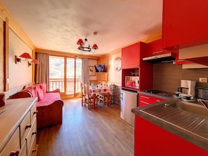 Una camera accogliente con pavimento in legno, un tavolo da pranzo con sedie, un divano, un angolo cottura con pensili rossi e una TV montata a parete.