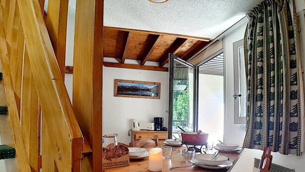 A dining area with a wooden table, chairs, a candle, and a magazine. - Charming duplex 6 pers, near slopes, north balcony, WiFi (Les Menuires)