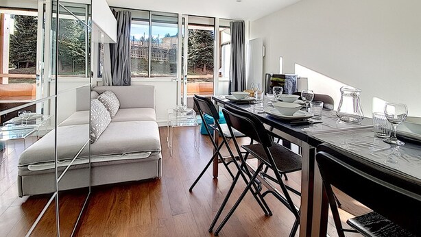 Ein modernes Wohnzimmer mit einer Couch, einem Esstisch und Stühlen. Große Fenster bieten einen Blick ins Freie.
