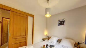 Een net opgemaakt bed met witte lakens, een nachtkastje met een lamp en een wandlamp.