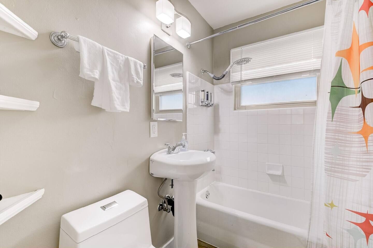 A bathroom with a white sink, toilet, and bathtub. There is a window with blinds and a shower curtain with a star pattern.