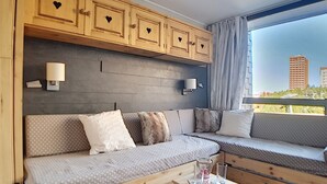 Ett modernt vardagsrum med en soffa i trä, ett litet träbord och utsikt över byggnader genom stora fönster.