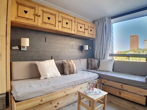 Ein modernes Wohnzimmer mit einer Holzcouch, einem kleinen Holztisch und Blick auf Gebäude durch große Fenster.