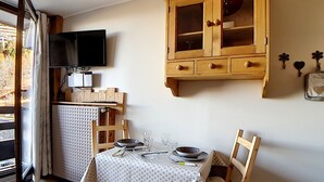 En matplats med ett bord dukat för två personer, trästolar, ett skåp med glasdörrar och en tv som är monterad på väggen.