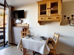 Ein Essbereich mit einem Tisch für zwei Personen, Holzstühlen, einem Schrank mit Glastüren und einem an der Wand montierten Fernseher.