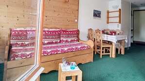 Ein gemütliches Zimmer mit einer Holzcouch, einem Esstisch mit Stühlen und einem kleinen Holzhochentisch mit einem Glas.