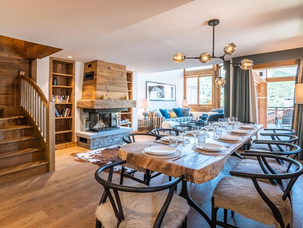 A spacious dining area with a long wooden table set for six, surrounded by chairs with fur throws. A stone fireplace with a wooden mantel is visible in the background. - Luxurious chalet, direct slope access, parking and WIFI included (Saint-Martin-de-Belleville)