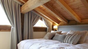 Quarto com teto de madeira, uma cama grande com várias almofadas e uma janela com cortinas.