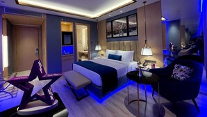 A modern hotel room with a large bed, bedside tables, a desk, and a seating area with a chair and bench. - Planet Hollywood (Thane)