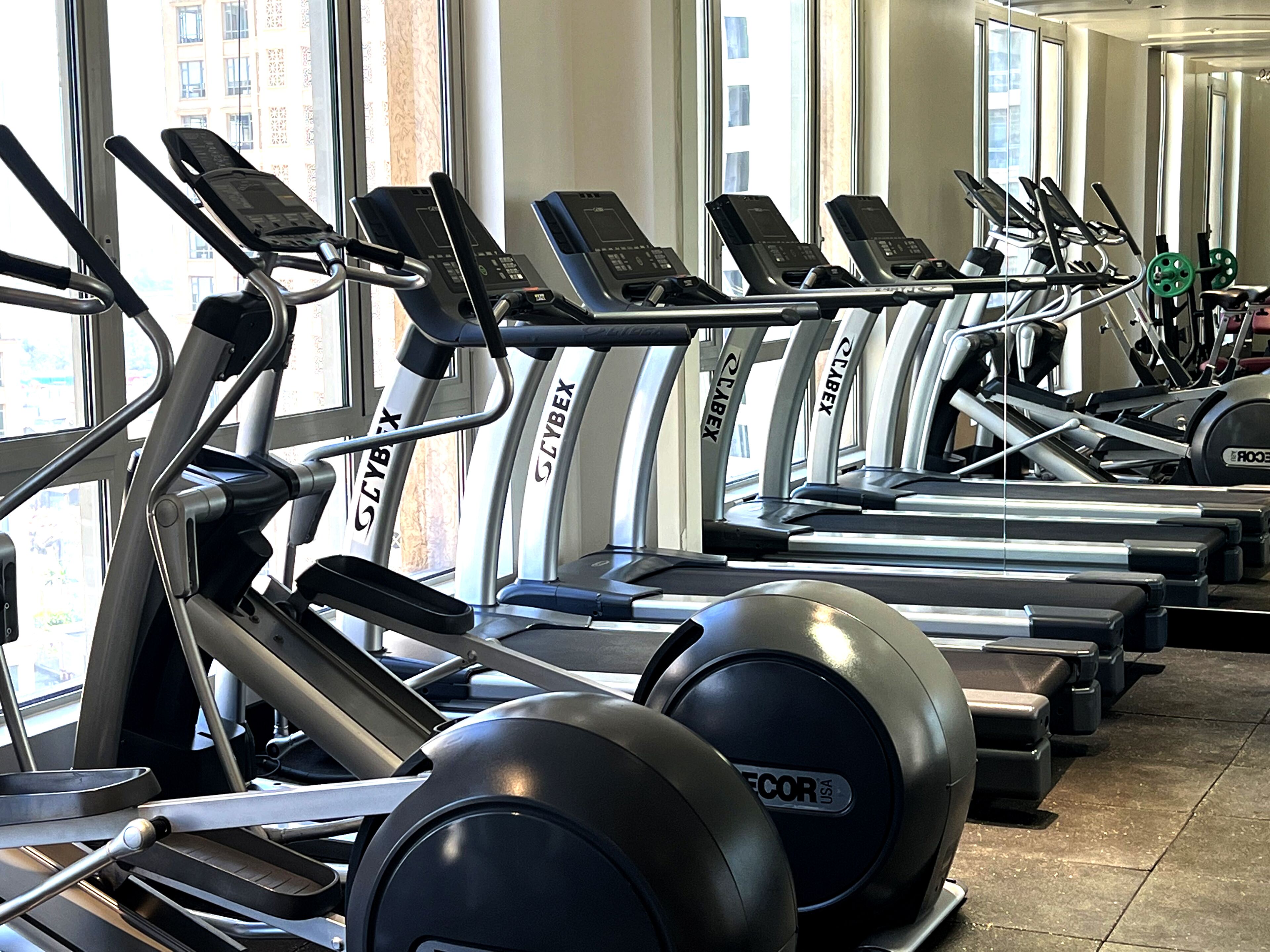 Eine Reihe von Laufbändern der Marke 'GYBEX', angeordnet in einem Fitnessstudio mit großen Fenstern und natürlichem Lichteinfall.