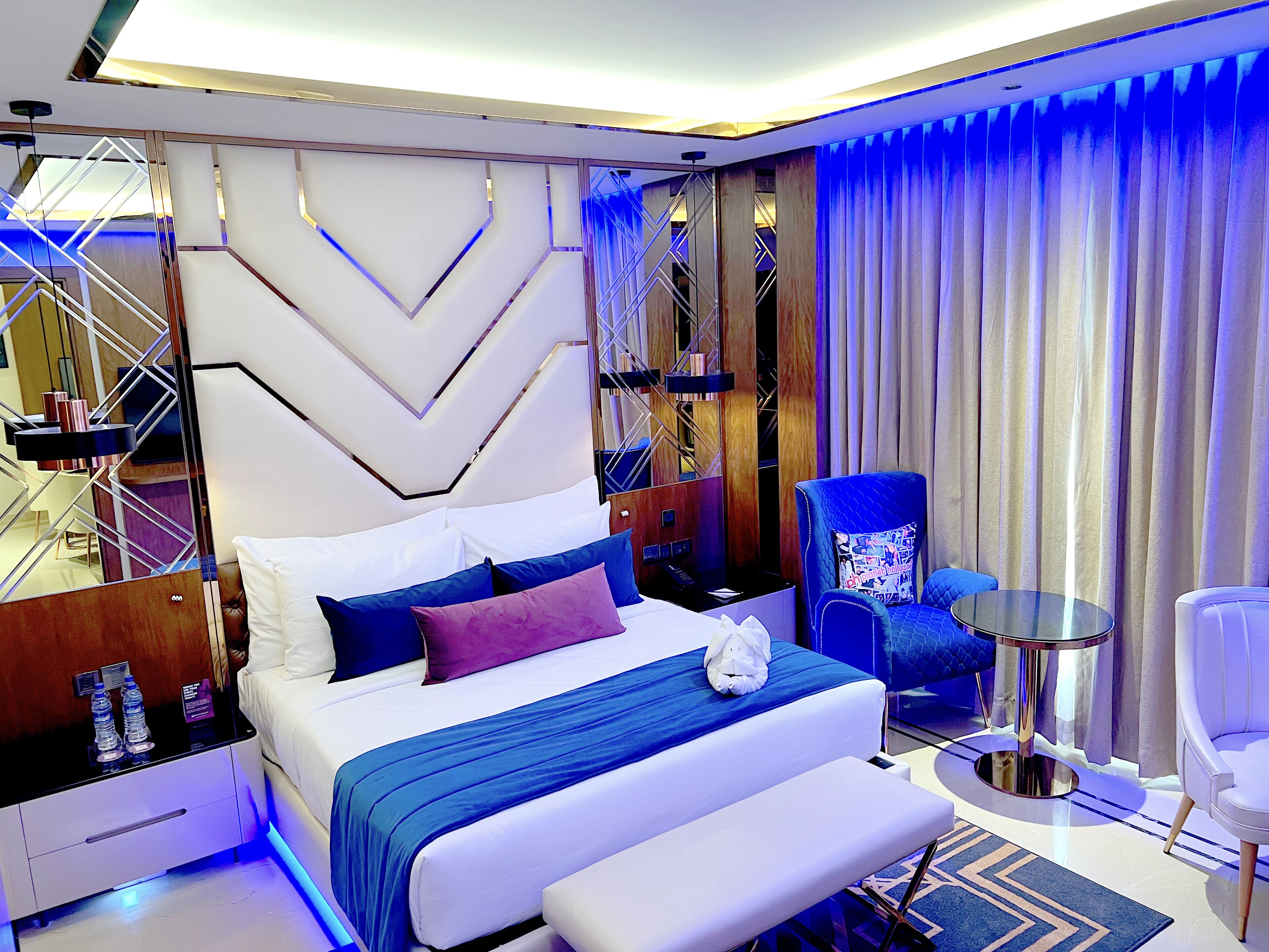 A modern hotel room with a large bed, a seating area with a chair and a table, and a view of the city through large windows.