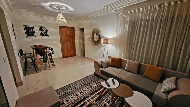 Un salon avec un canapé, une table basse et un tapis à motifs. On y trouve également un coin repas avec une table et des chaises, ainsi qu’une porte en bois.