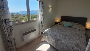 A bedroom with a large window, a bed with a patterned bedspread, a nightstand with a lamp, and a wall-mounted light fixture.