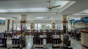 A spacious restaurant with high ceilings, multiple ceiling fans, and large windows offering a view of the outdoors. - Hotel Falcon Crest (Kandaghat)