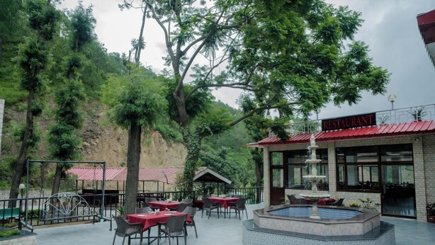 An outdoor restaurant with tables and chairs, a fountain, and a view of a forested hillside. - Hotel Falcon Crest (Kandaghat)