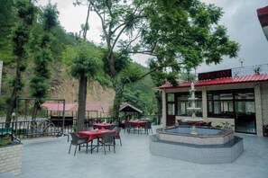 An outdoor restaurant with tables and chairs, a fountain, and a view of a forested hillside. - Hotel Falcon Crest (Kandaghat)