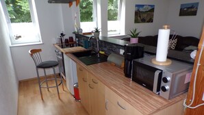 A compact kitchen with wooden cabinets, a built-in sink, a microwave, and a coffee maker. There are two windows providing natural light, and a small potted plant on the counter.