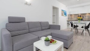 Ett modernt vardagsrum med en grå soffa, ett litet vitt bord med en växt och frukt på, och ett kök i bakgrunden med kakel på väggen ovanför arbetsbänken.